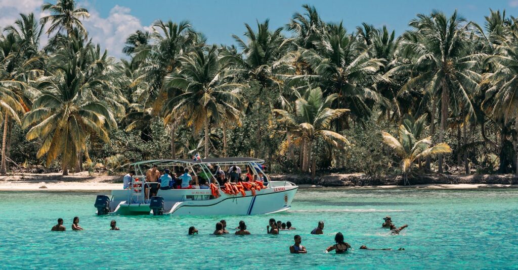 Sunny day at a tropical beach with people swimming and a boat near coconut palms.